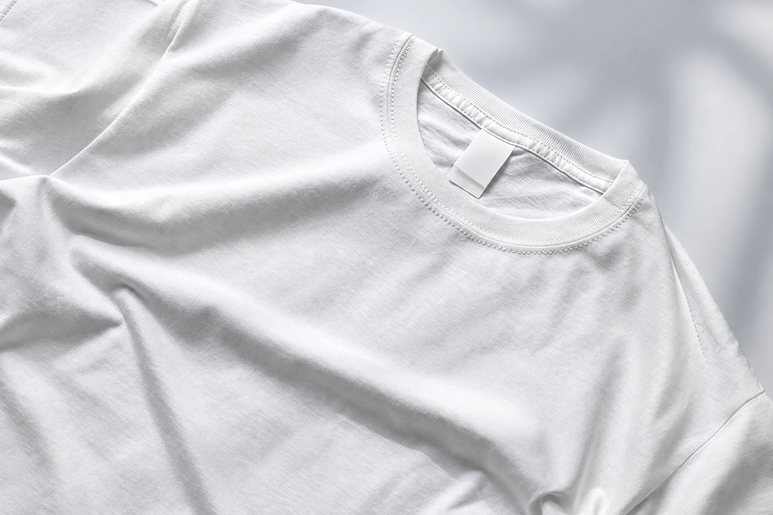 Close overhead view of a plain white crew-neck T-shirt laid flat on a light gray surface.