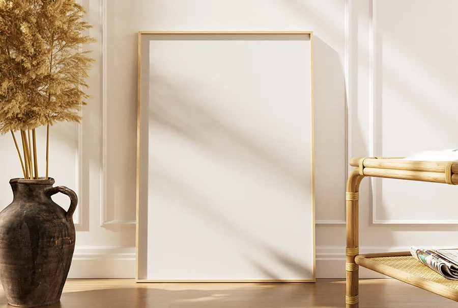 Blank light-wood framed poster leaning against a sunlit paneled wall, with a dark vase of dried foliage and a woven cane chair.