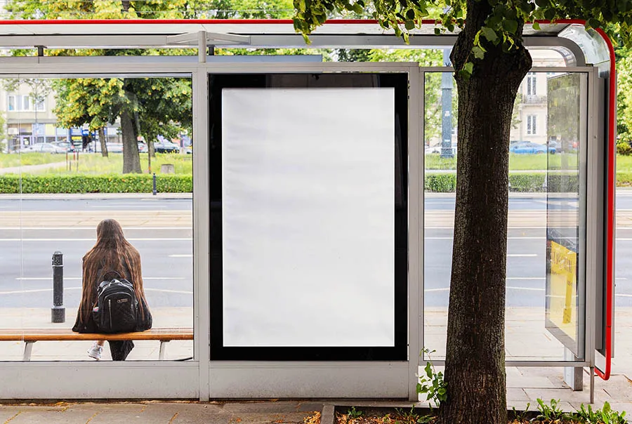 Blank white poster in a black-framed glass bus shelter beside a street, trees, and a seated passenger.