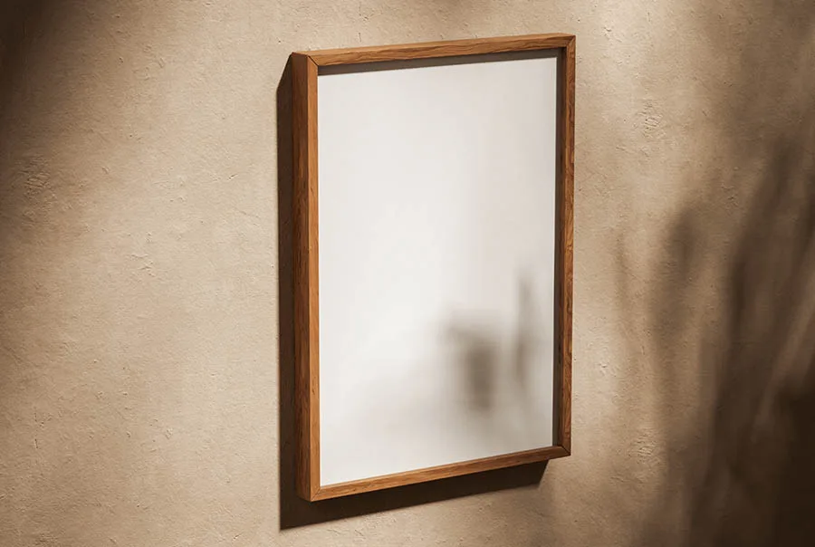 Portrait poster in a warm brown oak frame on a textured beige wall, lit from the side.