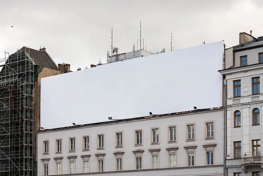 Blank rooftop billboard above a pale stone city building, with scaffolding at left and an overcast sky.