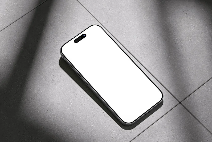 Angled iPhone with a blank white screen resting on sunlit gray tiles, casting a sharp shadow.