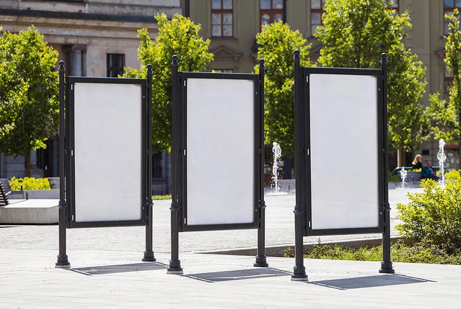 Three blank, black-framed citylight poster displays stand in a sunny plaza with trees and a stone building.