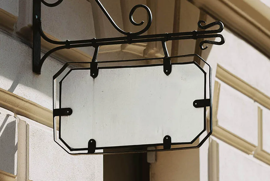 Blank pale sign panel with clipped corners and a dark border, hanging from an ornate black wrought-iron bracket on a sunlit exterior wall.
